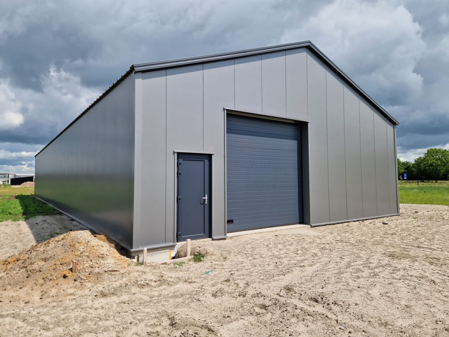 Grijze loods in Lemelerveld met zadeldak en gesloten sectionaalpoort onder een bewolkte hemel, op een zandvlakte